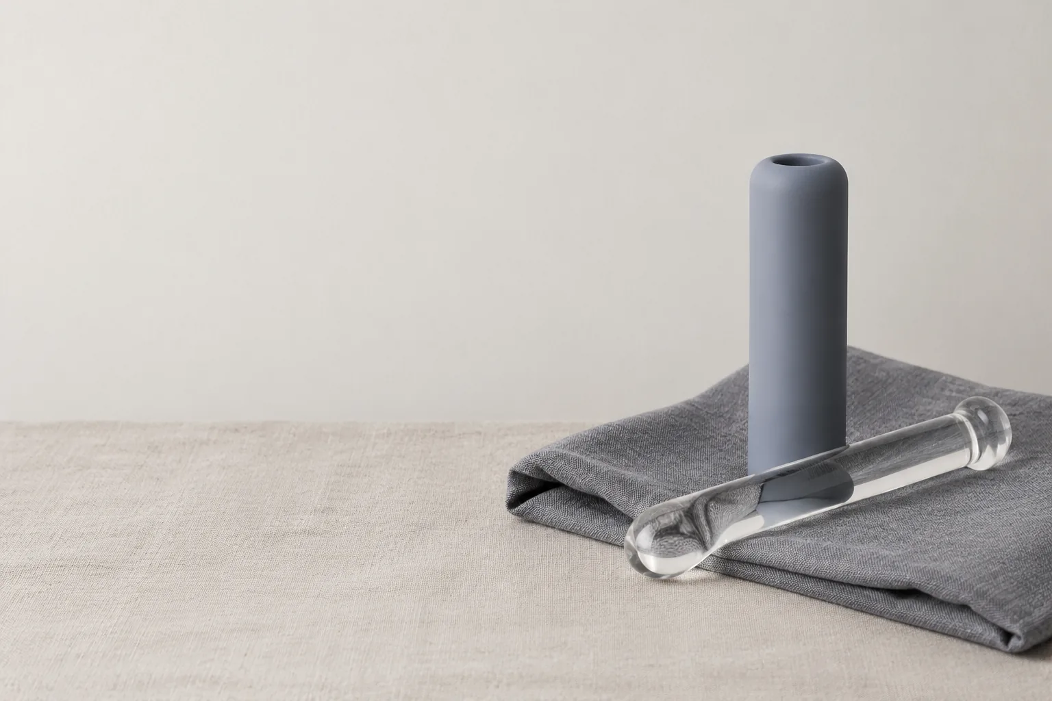 A still life on pale linen: a matte silicone cylinder and a clear glass rod resting on a folded stone-grey cloth, with the objects grouped to the right of frame